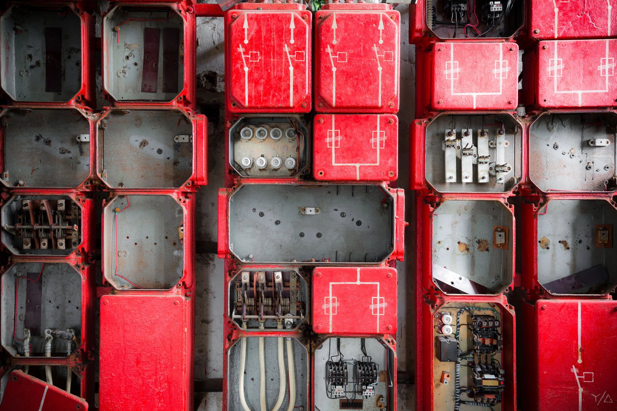 Ein alter, rostiger Sicherungskasten mit offenen Fächern und elektrischen Bauteilen. Die roten Metallrahmen sind abgenutzt, während freiliegende Kontakte und Korrosion den industriellen Verfall betonen.